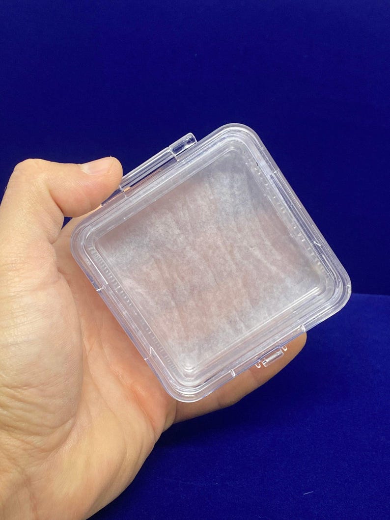 May include: A clear plastic square box with a hinged lid. The box is empty and has a white paper liner.