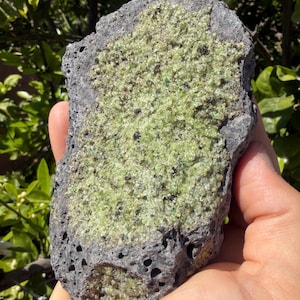 Pode incluir: Uma rocha cinza escura áspera com uma superfície cristalina verde clara. A rocha tem uma textura porosa com pequenos buracos e uma forma natural irregular. Os cristais verdes estão agrupados.