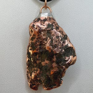 May include: A copper pendant with a unique, irregular shape, suspended from a black cord. The pendant has a textured surface with a mix of copper and darker tones, and a silver-colored metal cap at the top.