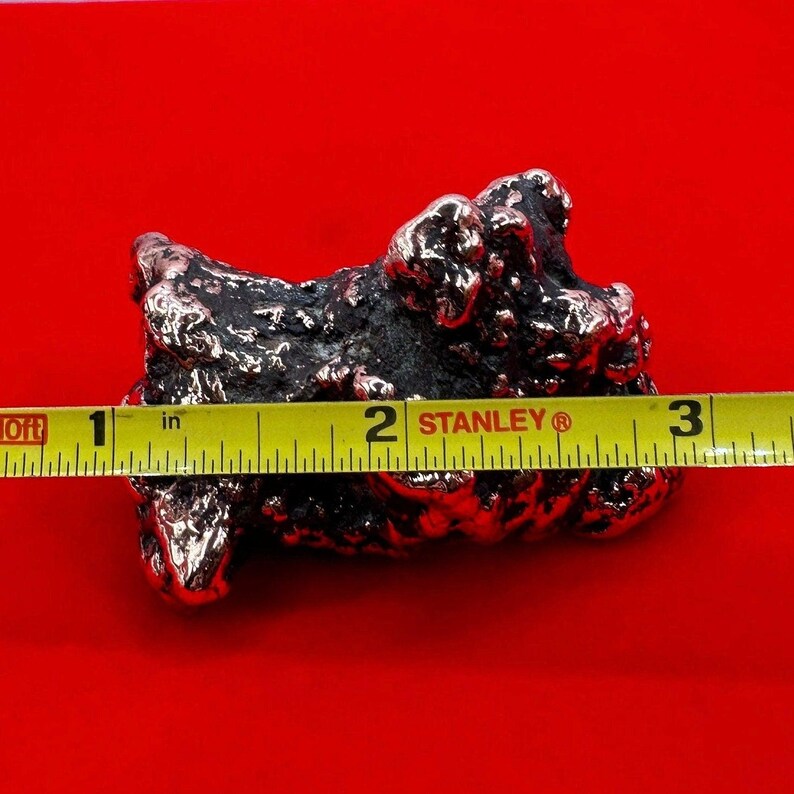 May include: A close-up of a rough, silver-colored rock with a textured surface. The rock is measured with a yellow tape measure, showing it is approximately 2 inches long.