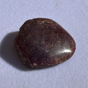 May include: A dark red, irregularly shaped gemstone with a smooth, polished surface.