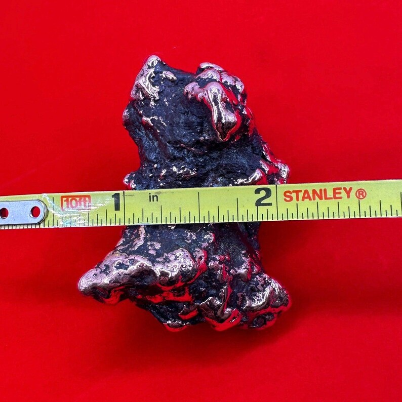 May include: A rough, irregular shaped piece of metal with a silver and copper finish. The metal is measured with a tape measure, showing it is about 2 inches long.