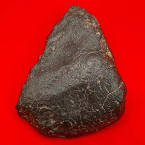 Pode incluir: Uma rocha escura de forma irregular, com uma superfície rugosa e texturizada e uma rede de finas rachaduras. A rocha é de cor carvão escuro e assenta sobre um fundo vermelho brilhante. A forma geral é triangular.
