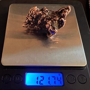May include: A rough, irregular, copper-colored rock on a digital scale. The scale reads 12.174 grams.