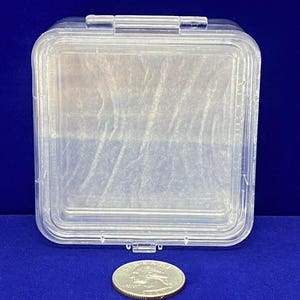 May include: A clear plastic square storage container with a hinged lid. The container is about the size of a UK 20 pence coin.