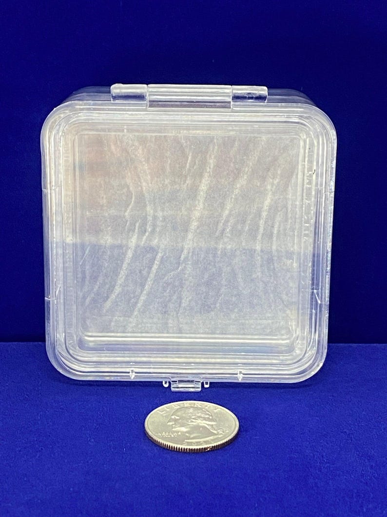 May include: A clear plastic square storage container with a hinged lid. The container is about the size of a UK 20 pence coin.