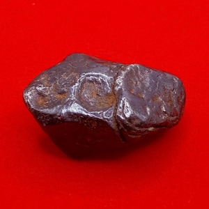 May include: A dark brown, metallic-looking rock with a smooth, polished surface and irregular, pitted indentations. The object has a dense, heavy appearance with visible rust-coloured oxidation within the deeper crevices of its textured exterior.