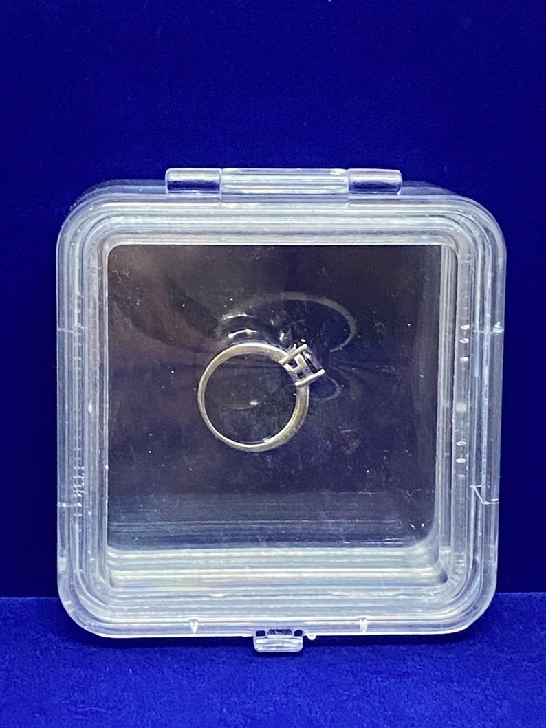 May include: A clear plastic ring box containing a silver ring with a single clear stone.