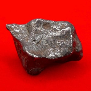 May include: A metallic gray, irregularly shaped meteorite against a red background. The surface of the space rock is textured with ridges and indentations, showcasing a rugged, natural appearance. The object appears to be a solid, dense mass.