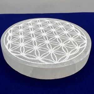 May include: A white selenite charging plate with a carved flower of life design. The plate is round and has a smooth, polished surface.