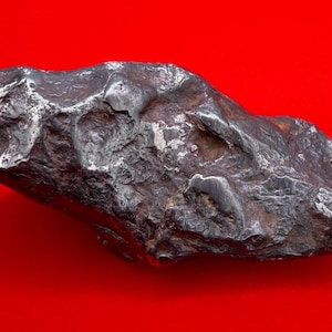 Puede incluir: Un espécimen de meteorito metálico de color gris oscuro con una textura superficial rugosa e irregular. El objeto presenta hendiduras profundas, áreas pulidas suaves y bordes afilados en toda su forma, mostrando una apariencia densa y pesada.