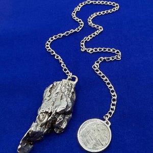 May include: A silver-colored metal pendant with a rough, irregular, rock-like texture attached to a silver-colored chain. The chain also features a small, round, flat silver-colored coin-shaped charm with a textured surface and a visible etched design.