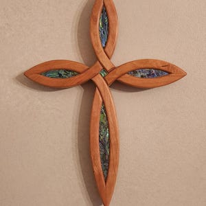 May include: Wooden cross with a unique design. The cross is made of light brown wood and features inlaid sections of iridescent abalone shell. The cross is mounted on a neutral-colored wall.