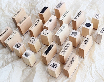 May include: A selection of wooden stamps with diverse designs and text. The stamps display geometric patterns, floral motifs, and phrases. The stamps are arranged on a light brown, textured surface.
