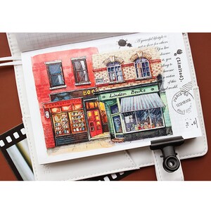 May include: Watercolor illustration of a red brick building with two storefronts. The storefront on the left says "The Bookworm Company" and the storefront on the right says "Lindam Books".