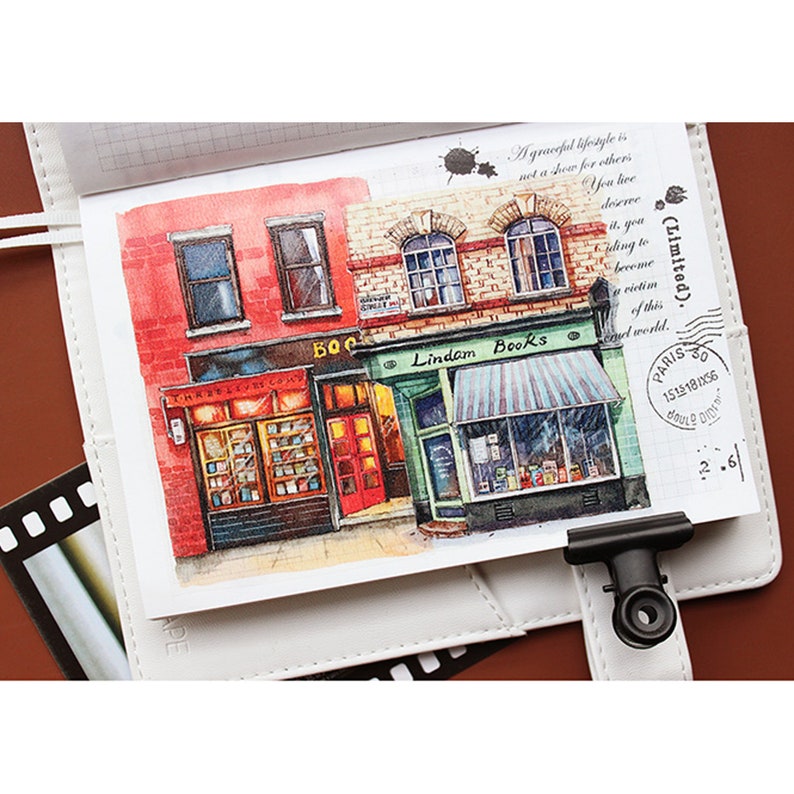 May include: Watercolor illustration of a red brick building with two storefronts. The storefront on the left says "The Bookworm Company" and the storefront on the right says "Lindam Books".