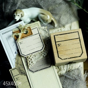 May include: A wooden stamp with a rectangular design and the number "No." printed on it, alongside vintage-style tags and a ceramic dog figurine. The tags have a distressed look with the text "No." and lines for writing. The image also shows a piece of lace and the dimensions 45x40cm.