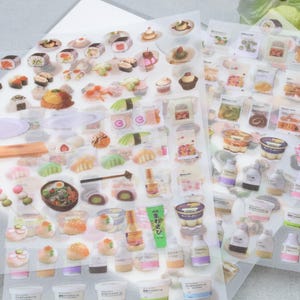 May include: Sheets of stickers featuring illustrations of various Japanese foods, including sushi, desserts, and packaged items. The stickers are colorful and detailed, with a variety of shapes and sizes. The stickers are on a clear backing.