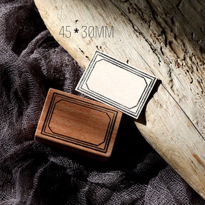 May include: Two wooden stamps with a black and white rectangular frame design. The stamps are 45 x 30 mm. One stamp is on a wooden surface and the other is on a grey fabric surface.