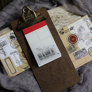 K&ouml;nnte beinhalten: Eine braune Klemmbrett mit einem wei&szlig;en Papierblock befestigt. Der Papierblock hat ein Gittermuster und einen Stempel mit B&auml;umen. Das Klemmbrett hat einen roten und goldenen Clip. Das Klemmbrett befindet sich auf einer braunen Holzoberfl&auml;che mit einem St&uuml;ck grauem Stoff.