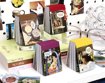 May include: A collection of small, colorful notebooks with various images and text on the covers. The notebooks have different colored covers, including brown, red, purple, and yellow. The image also includes other decorative items like photos and a clear glass object.