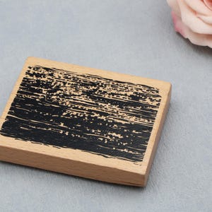 May include: A rectangular wooden block stamp featuring a black ink design on its top surface. The design consists of horizontal, distressed, and textured black brushstrokes that cover the majority of the light-colored wooden surface area.