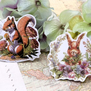 May include: Two die-cut stickers featuring woodland creatures. One shows a squirrel with a fluffy tail, holding flowers. The other depicts a rabbit surrounded by a floral wreath. Both stickers have a vintage, botanical illustration style.