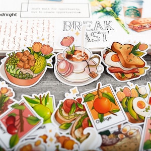 May include: A selection of vibrant stickers with breakfast-themed designs. The stickers depict a bowl of food, a teacup with macarons, a sandwich, and various fruits. The word "BREAKFAST" is displayed in the background.