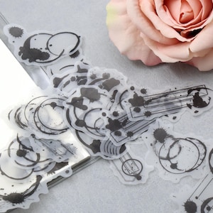 May include: A collection of transparent stickers with black ink blot and geometric designs. The stickers are scattered on a white surface, with a soft pink rose in the background. The stickers feature circles, lines, and ink splatters.