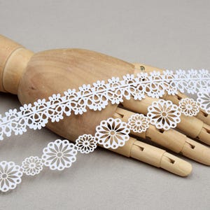 May include: Two white lace ribbons with floral and scalloped designs are displayed on a wooden hand. The lace features intricate cut-out patterns, with one ribbon showcasing a daisy-like motif. The background is a neutral gray.