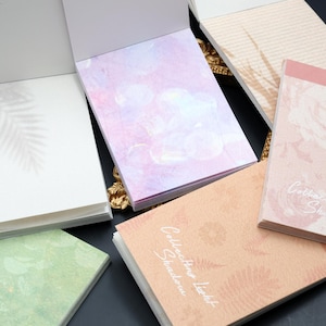 May include: A collection of decorative notepads with floral and botanical designs. The notepads feature various colors, including pink, green, and peach, with printed text. Some pads have open pages, revealing the blank interior.