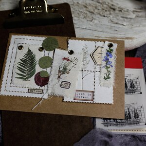 K&ouml;nnte beinhalten: Ein braunes Papierjournal mit verschiedenen gepressten Pflanzen und Ephemera, die an den Seiten befestigt sind. Das Journal hat einen wei&szlig;en Hintergrund und der Text "Lost in Forest" ist auf einer der Seiten gedruckt.