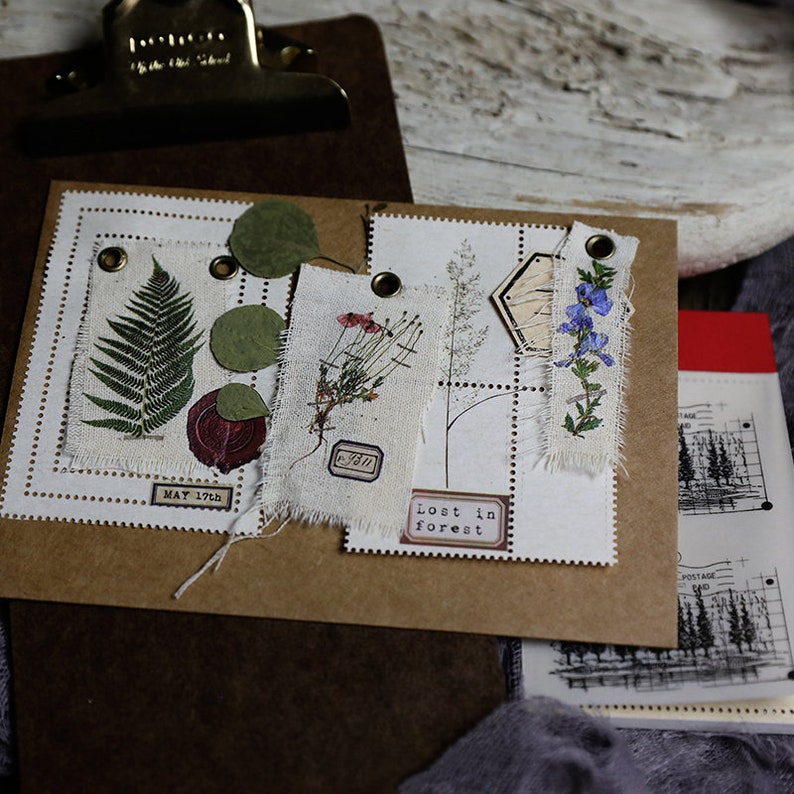 K&ouml;nnte beinhalten: Ein braunes Papierjournal mit verschiedenen gepressten Pflanzen und Ephemera, die an den Seiten befestigt sind. Das Journal hat einen wei&szlig;en Hintergrund und der Text "Lost in Forest" ist auf einer der Seiten gedruckt.