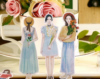 May include: Three watercolor illustrations of women wearing dresses and hats. The women are holding flowers. The illustrations are printed on white paper and are ready to be cut out and used for scrapbooking or other craft projects.