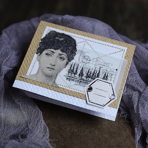 K&ouml;nnte beinhalten: Eine handgemachte Karte mit Vintage-&Auml;sthetik. Die Karte zeigt ein Schwarzwei&szlig;bild einer Frau mit einem dekorativen Rand. Die Karte enth&auml;lt auch ein Schwarzwei&szlig;bild eines Waldes mit dem Text "POSTAGE PAID" und ein leeres Etikett mit zwei Metall&ouml;sen.