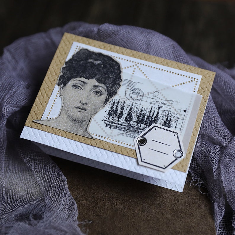 K&ouml;nnte beinhalten: Eine handgemachte Karte mit Vintage-&Auml;sthetik. Die Karte zeigt ein Schwarzwei&szlig;bild einer Frau mit einem dekorativen Rand. Die Karte enth&auml;lt auch ein Schwarzwei&szlig;bild eines Waldes mit dem Text "POSTAGE PAID" und ein leeres Etikett mit zwei Metall&ouml;sen.