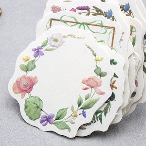 May include: A stack of scalloped-edge paper tags with watercolor floral designs. Each tag features a white background with a wreath of pink, purple, and yellow flowers and green leaves. A gold-colored decorative frame is in the background.