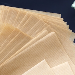 May include: A stack of light brown, rectangular parchment paper sheets, fanned out to display their textured, ribbed surface. The paper is arranged on a dark surface, with a hint of green and white foliage in the background.