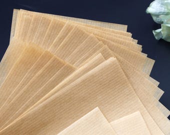 Może przedstawiać: Stos jasnobrązowych, prostokątnych arkuszy papieru pergaminowego, wachlarzowato ułożonych, aby pokazać ich teksturowaną, żebrowaną powierzchnię. Papier ułożony jest na ciemnej powierzchni, z odrobiną zielonych i białych liści w tle.