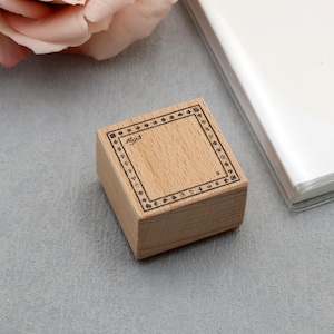 May include: Wooden stamp with a square design and a dotted border. The stamp has the word "Rgs" in the top left corner. A white notebook and a pink rose are in the background. The stamp is on a grey surface.