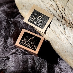 May include: A wooden stamp with the text "A World of Hanks" in a handwritten style, next to a matching stamp on paper. The stamps are on a dark grey fabric and a piece of wood.