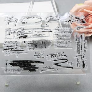 May include: A clear silicone stamp sheet featuring various black ink designs, including handwritten cursive script, typed text fragments, abstract brush strokes, and ink splatters. The designs include phrases like "Dear," "Longing," "Petrichor," and "Echoes of a forgotten telegram."