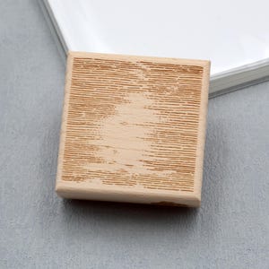 Op de afbeelding: Een kleine, rechthoekige houten stempel met een lichtbruine afwerking. Het oppervlak van de stempel heeft een gedetailleerd, getextureerd ontwerp van horizontale lijnen. De stempel ligt op een grijze ondergrond, met een wit object op de achtergrond.