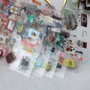 May include: Clear sheets of stickers with various illustrations, including cameras, coffee cups, and desserts. The stickers have a transparent background and are held by a person's hand. Some stickers have text, such as "c'est la vie" and "kindness is better".