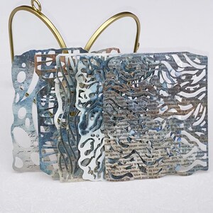 May include: An abstract paper art piece featuring layered, laser-cut designs in shades of blue, gray, and white. The artwork includes text elements and is held by a gold-colored metal frame.