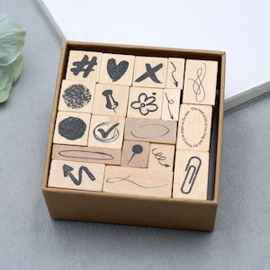 May include: A collection of wooden stamps featuring diverse symbols and shapes, such as a heart, tick, and paperclip, presented in a cardboard box. The stamps display black ink designs and are set on a grey surface.