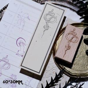 May include: A rectangular bookmark and a wooden stamp, both featuring a stylized snake design. The bookmark is cream-colored with a dark gray border. The stamp is on a wooden block. The text "60*30MM" is visible.