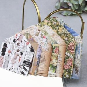 May include: A collection of decorative paper envelopes with vintage-style designs, including floral patterns, botanical illustrations, and fashion sketches. The envelopes are displayed in a gold-colored metal holder, creating a charming and artistic presentation.