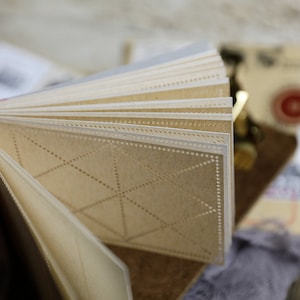 May include: A close-up of a small, cream-colored notebook with a geometric pattern on the pages. The pages are perforated and have a dotted border.