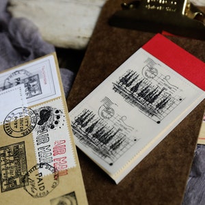K&ouml;nnte beinhalten: Zwei wei&szlig;e Papierstempel mit schwarzen Tintenzeichnungen von B&auml;umen und den Worten "Postage Paid" auf braunem Hintergrund.